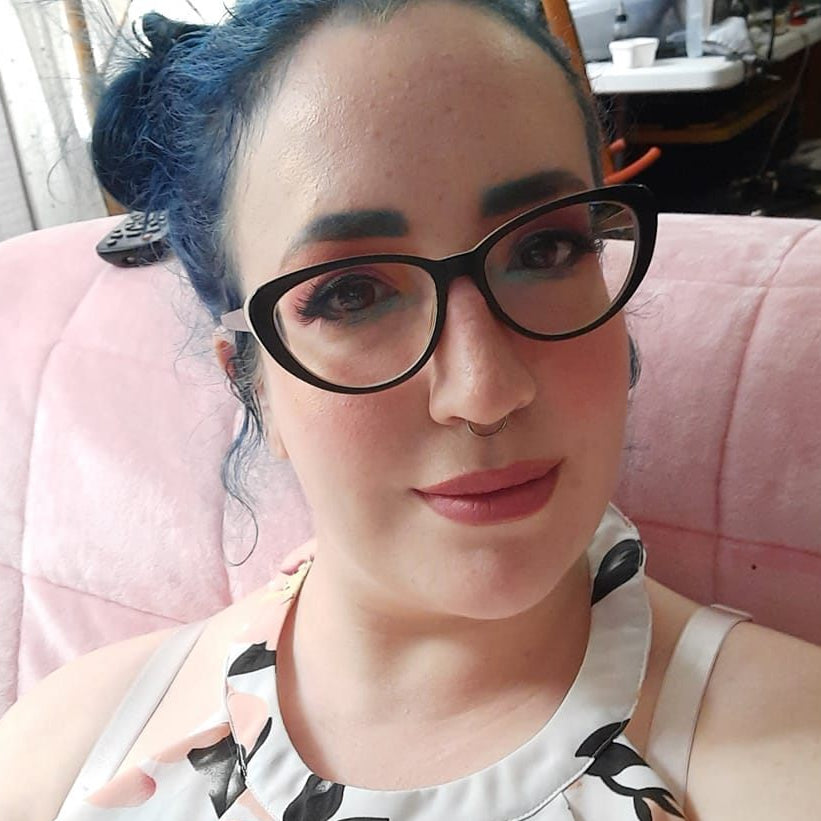 Person with blue hair and glasses sitting on a pink couch.